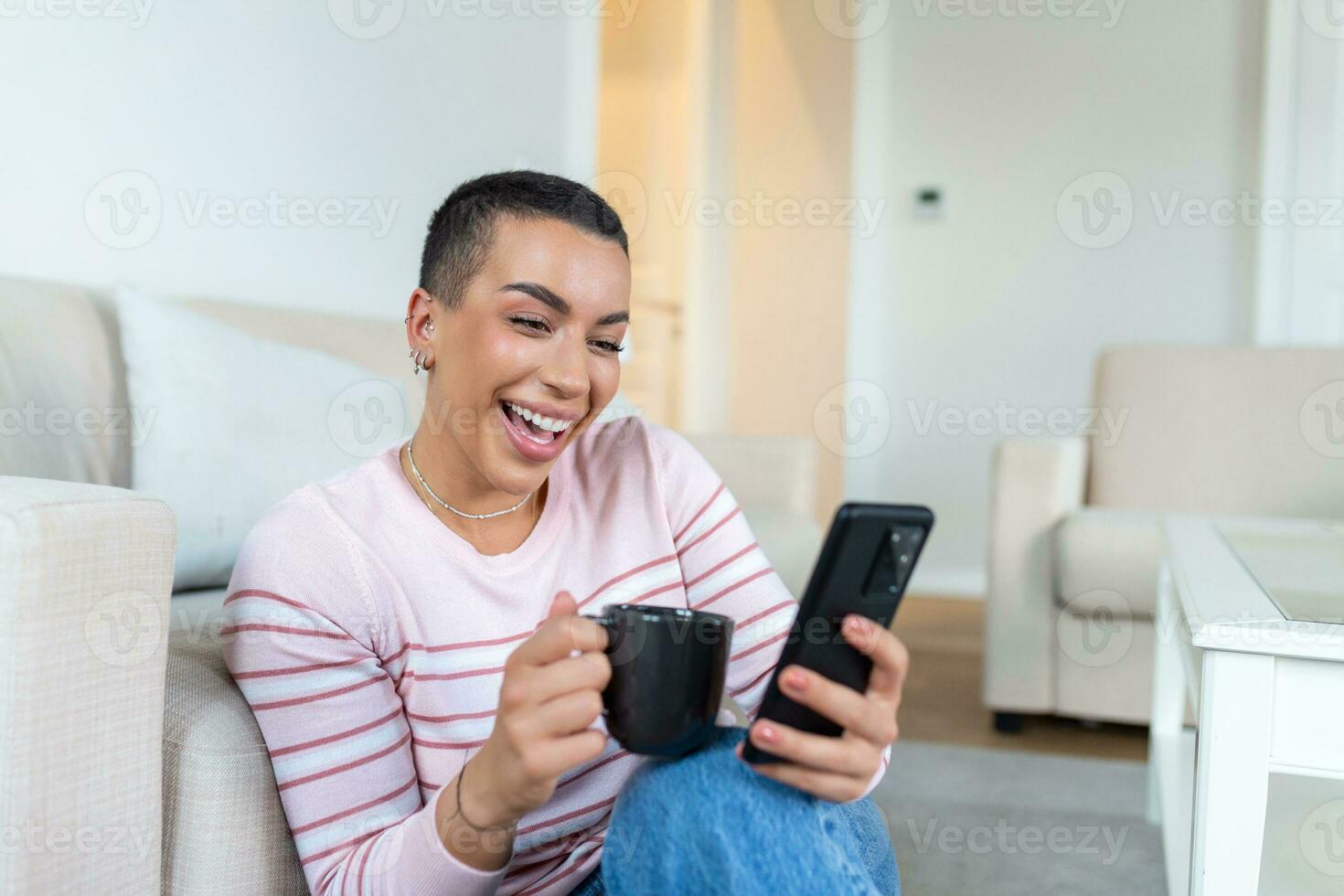 A young woman in a sweater with a cup in her hands looks at the phone while sitting on the couch. photo