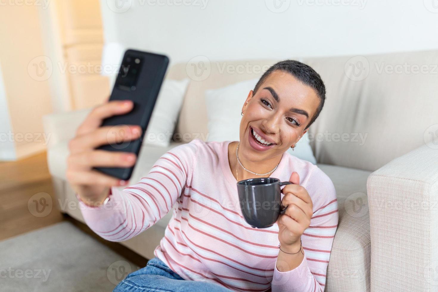 A young woman in a sweater with a cup in her hands looks at the phone while sitting on the couch. photo