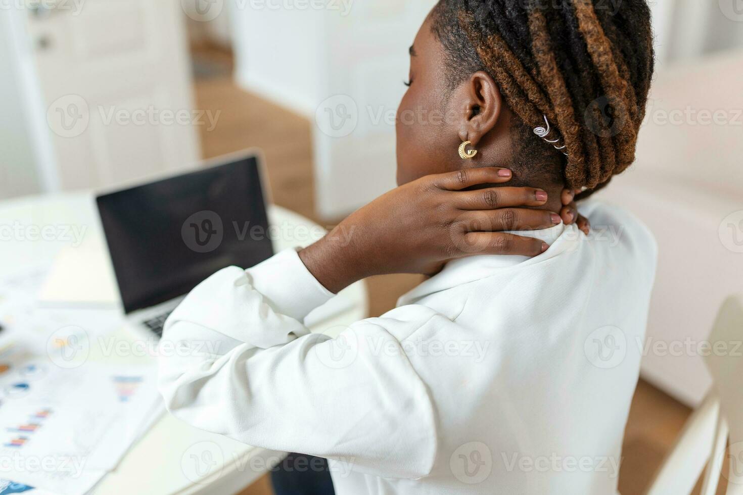 Portrait of young stressed African woman sitting at home office desk in front of laptop, touching aching shoulder with pained expression, suffering from shoulder ache after working on pc photo