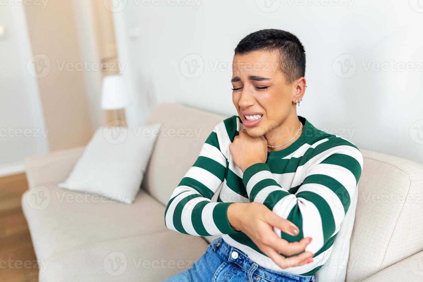 Young woman is sitting at home on her sofa and touching the her elbow while suffering from elbow pain photo