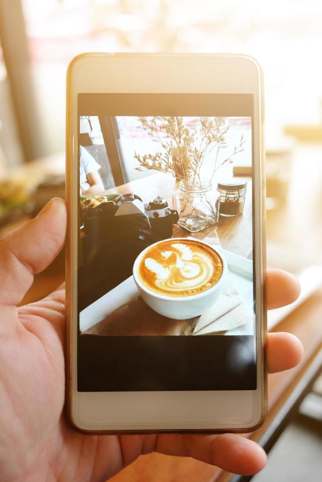 Beautiful hand Holding smartphone take a photo and screen shot Hot latte art coffee with vintage film camera and dry flower in grass vase with natural sulight in cafe