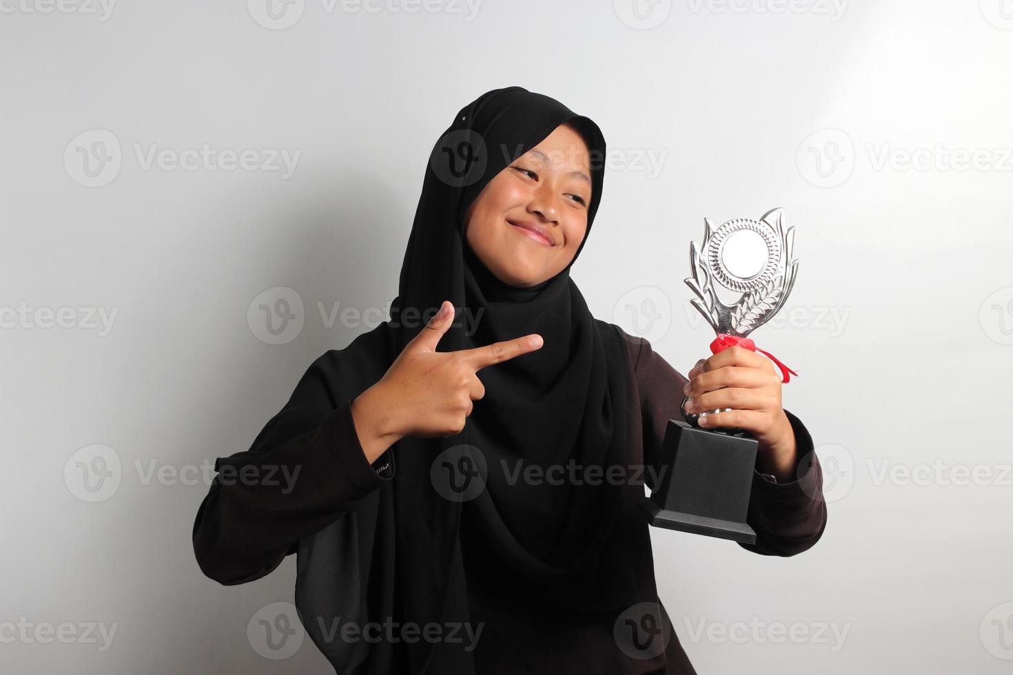 inteligente joven asiático niña estudiante en hijab señalando su trofeo aislado en blanco antecedentes. foto