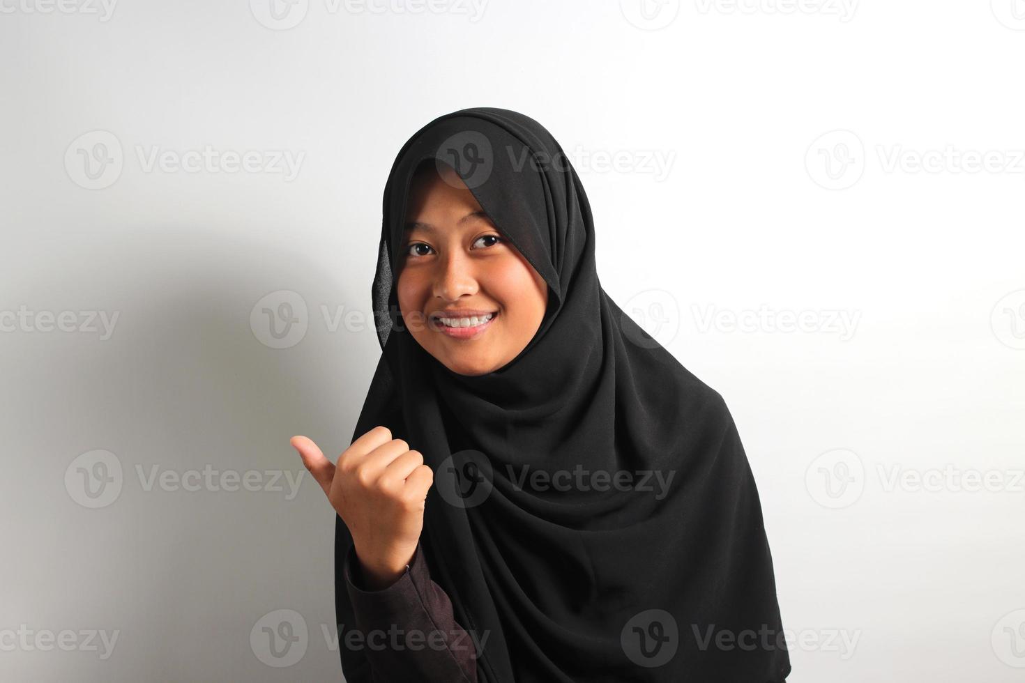 Cheerful young Asian girl in hijab pointing to the side with thumbs up isolated on white background. photo