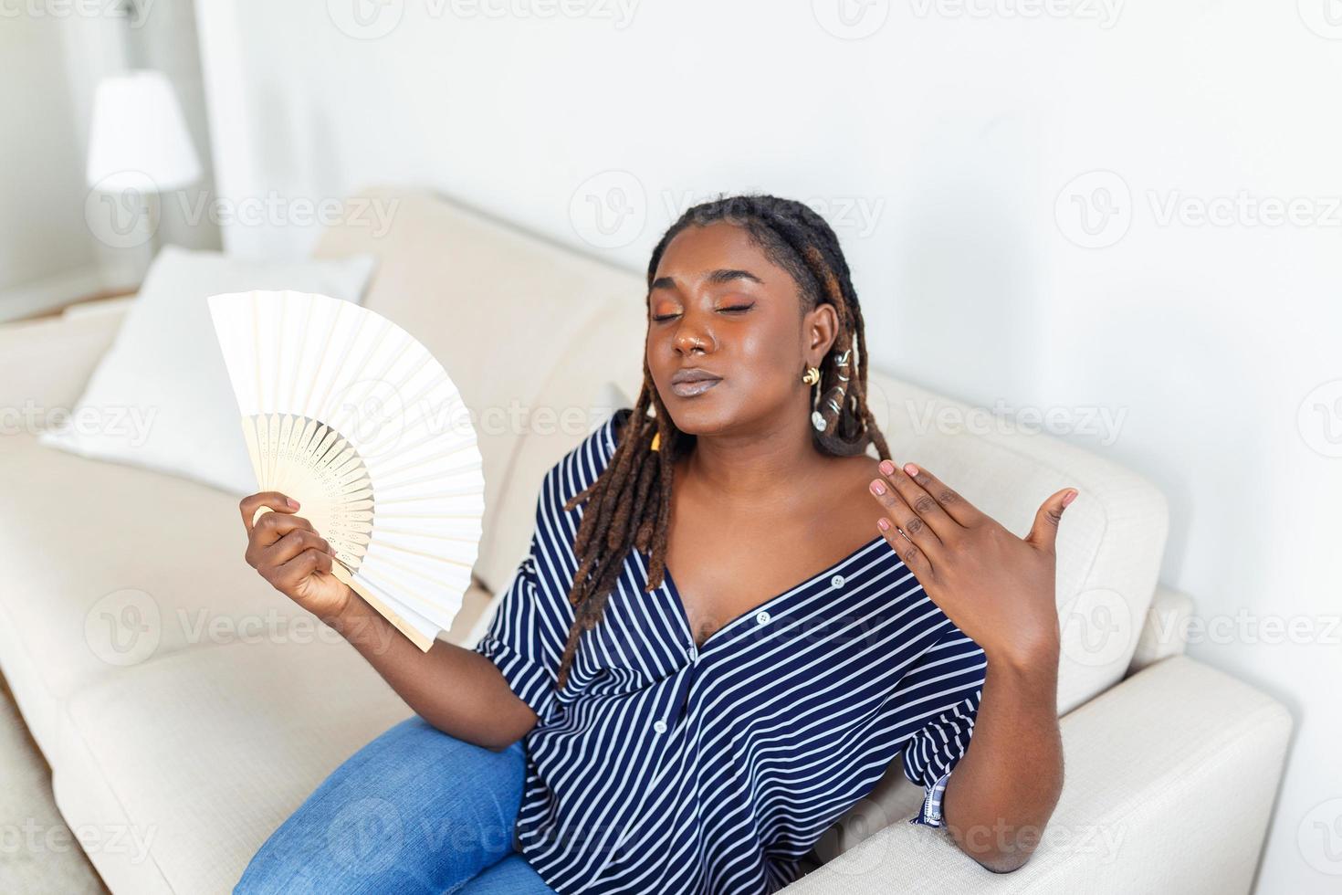 Overheated African woman sit on couch at home feel warm waving with hand fan cooling down, sweating girl relax on sofa in living room hold waver suffer from heat, no air conditioner system photo