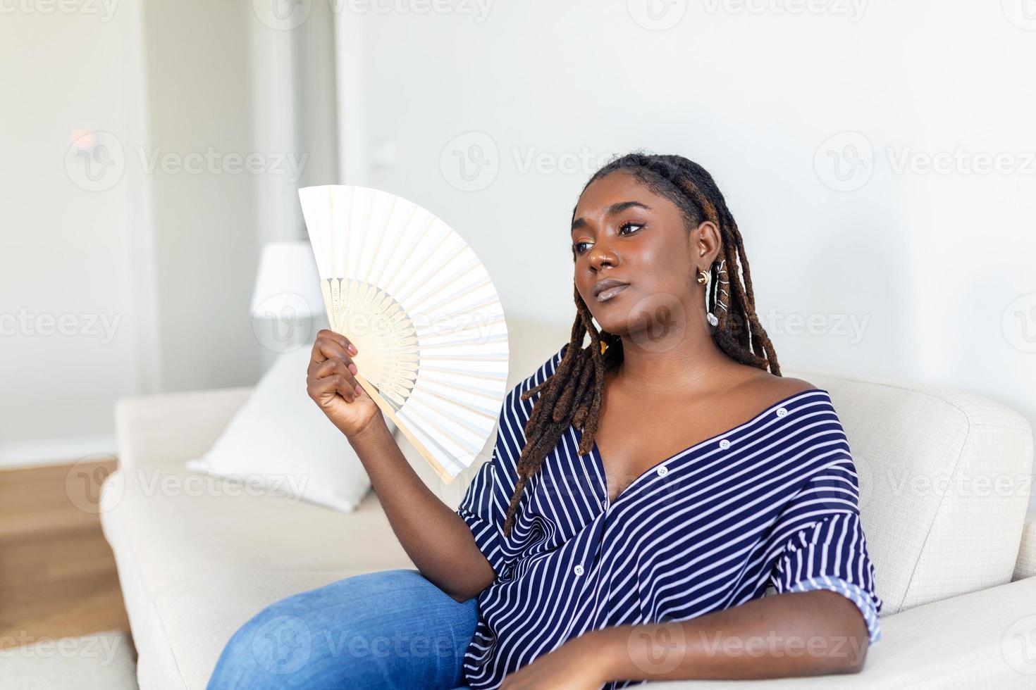 cansado afro mujer sufrimiento desde golpe de calor o caliente verano plano sin aire acondicionado, utilizando ondulación admirador, sentado en vivo habitación a hogar. calentamiento excesivo foto