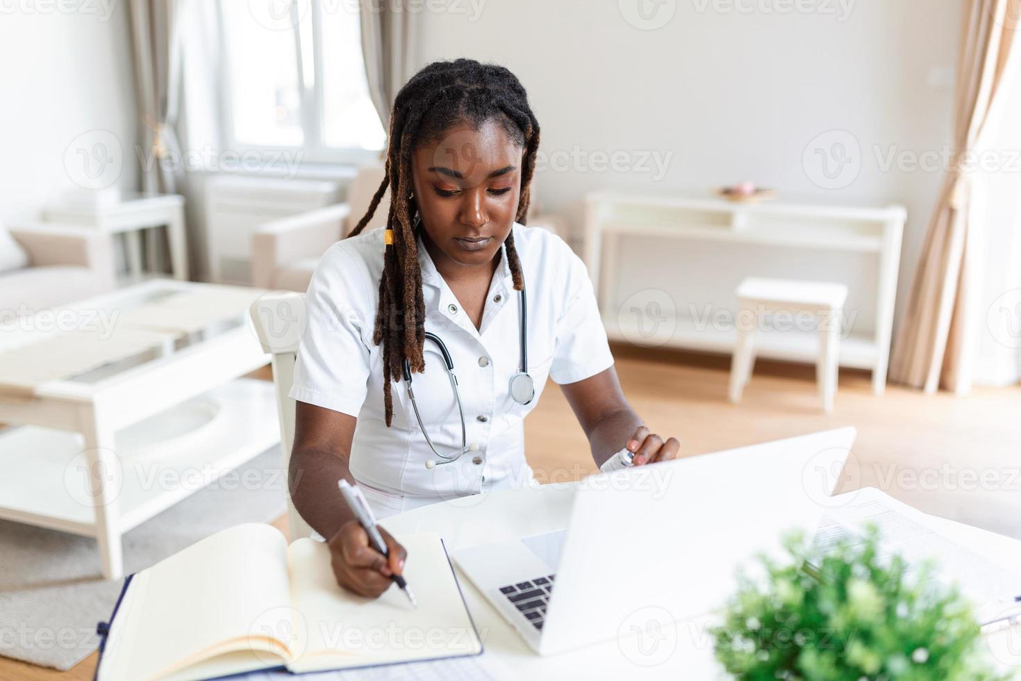 Smiling professional female doctor wearing uniform taking notes in medical journal, filling documents, patient illness history, looking at laptop screen, student watching webinar photo