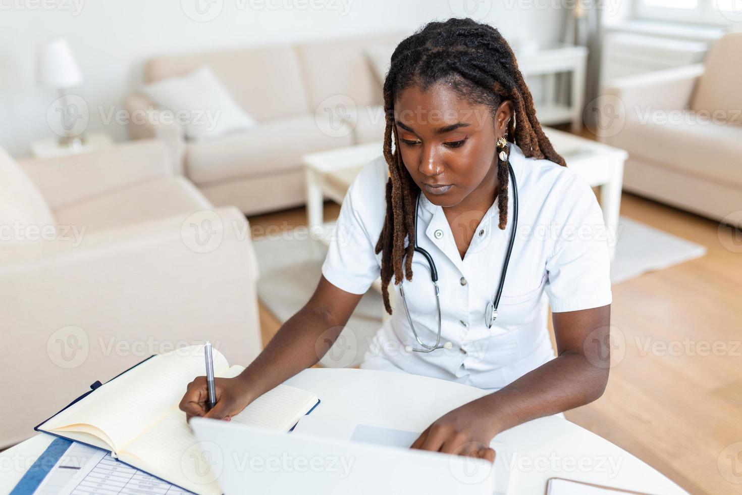 Smiling professional female doctor wearing uniform taking notes in medical journal, filling documents, patient illness history, looking at laptop screen, student watching webinar photo