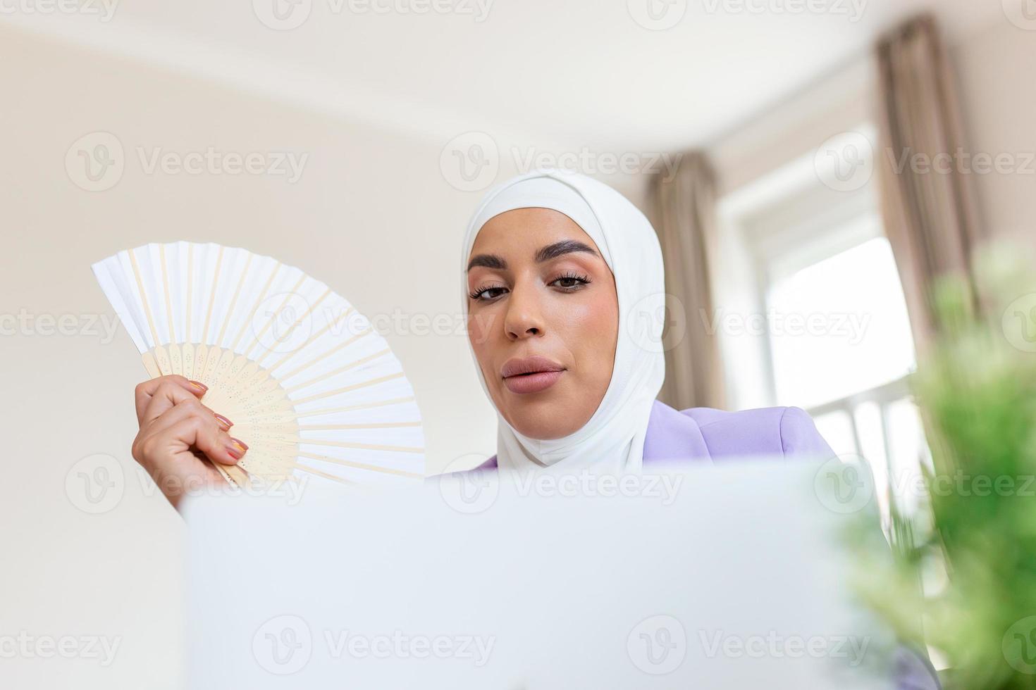 Young Arabic muslim woman hold wave fan suffer from heat sweating indoor work on laptop at home office, annoyed girl feel uncomfortable hot summer weather problem no air conditioner concept photo