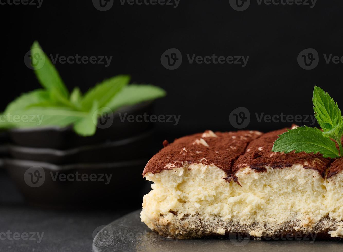 A piece of tiramisu sprinkled with cocoa, on top of a sprig of fresh mint on a black background photo