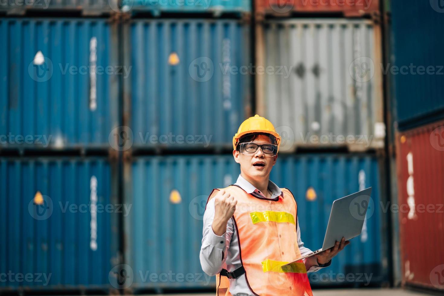 Asian technician dock worker in protective safety jumpsuit uniform and with hardhat and use laptop computer at cargo container shipping warehouse. transportation import,export logistic industrial photo