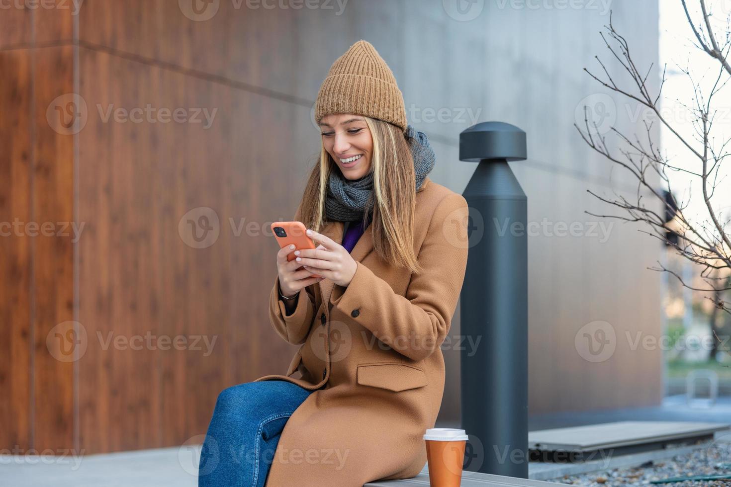 Image of beautiful stylish woman sitting on street bench with legs crossed on sunny day and holding mobile phone photo