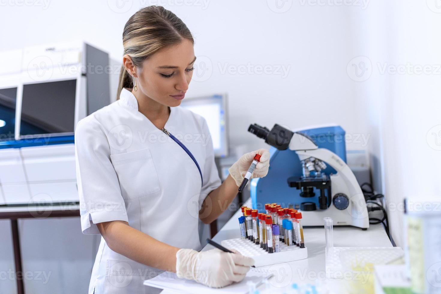Closeup portrait of woman scientist working in lab. Female researcher sitting at desk looking through microscope at blood and chemical test sample and making notes in clipboard. Biochemistry work photo