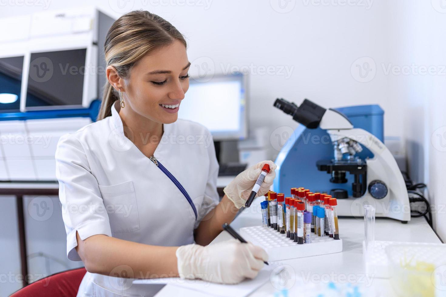 Closeup portrait of woman scientist working in lab. Female researcher sitting at desk looking through microscope at blood and chemical test sample and making notes in clipboard. Biochemistry work photo
