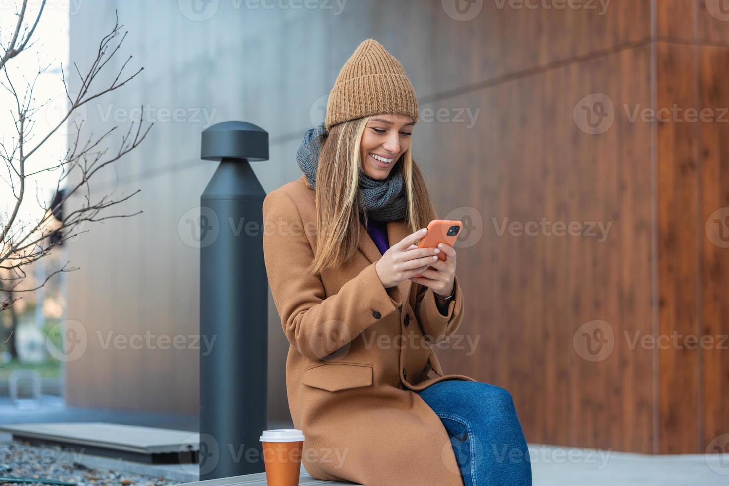 Image of beautiful stylish woman sitting on street bench with legs crossed on sunny day and holding mobile phone photo