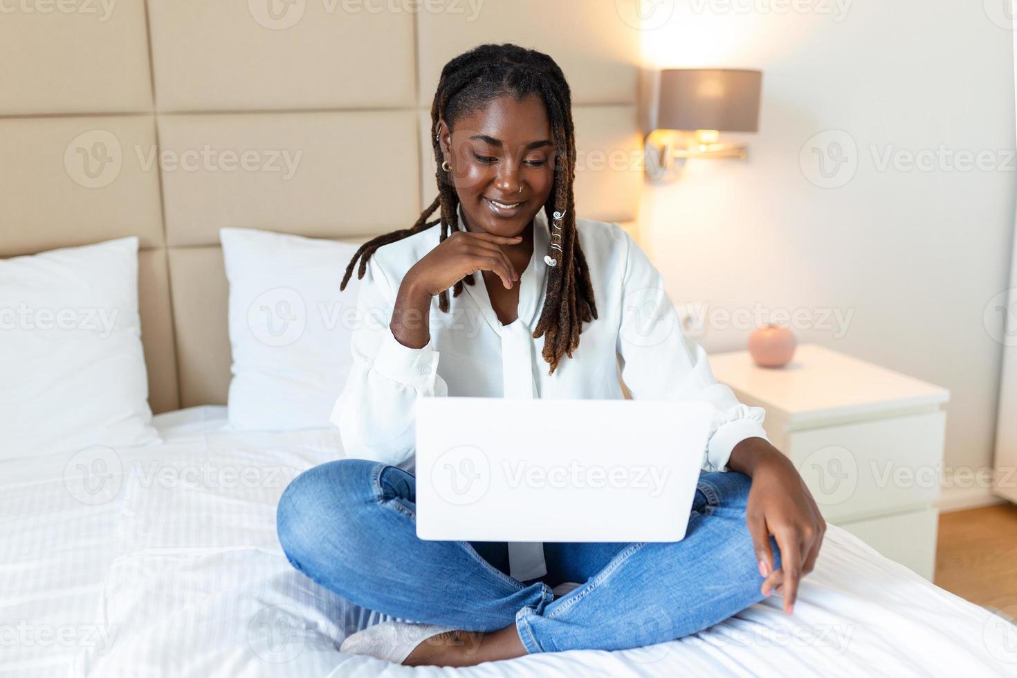 Attractive African woman in white shirt using laptop and smiling at morning. Video chat and freelance working from home concept of beautiful young woman with laptop in bed photo