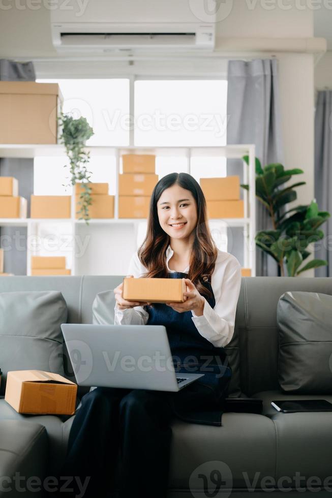 joven asiático negocio propietario mujer preparar paquete o empaquetar caja y en pie cheque en línea pedidos para entregar a cliente en tableta, ordenador portátil compras en línea concepto. foto