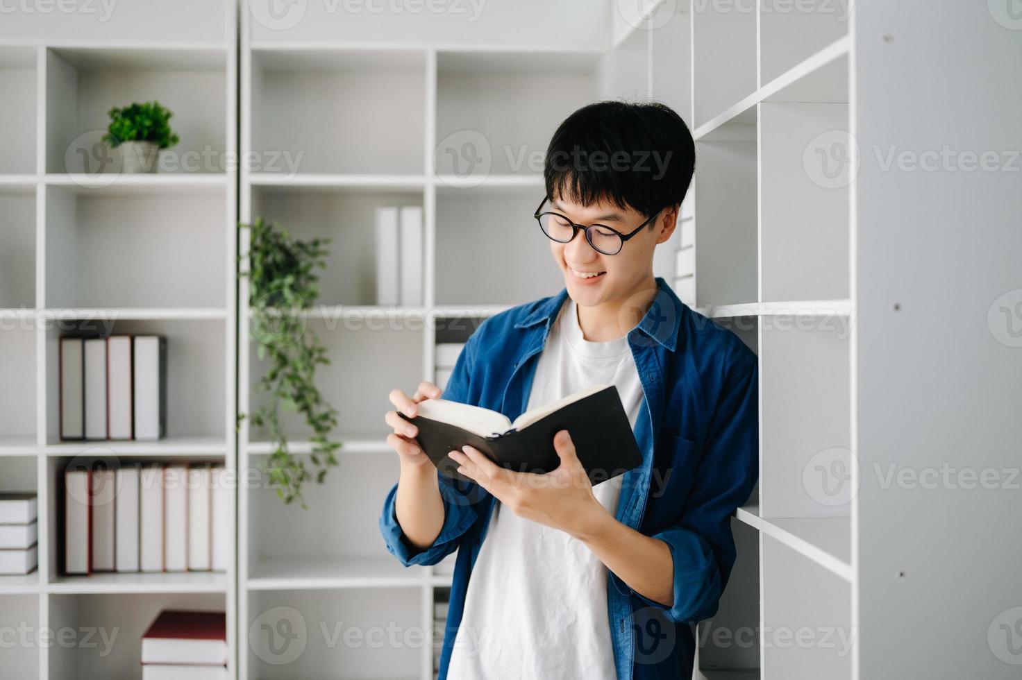 masculino estudiante tomando notas desde un libro a biblioteca, joven asiático sentado a escritorio haciendo asignaciones en Universidad biblioteca foto