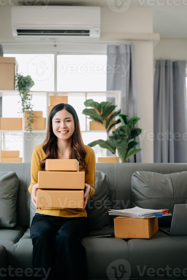 joven asiático negocio propietario mujer preparar paquete o empaquetar caja y en pie cheque en línea pedidos para entregar a cliente en tableta, ordenador portátil compras en línea concepto. foto