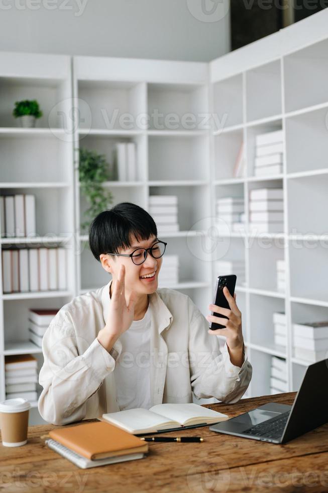 Attractive happy young Asian student studying at the college library, sitting at the desk, using a laptop, tablet and headphones having a video chat. photo