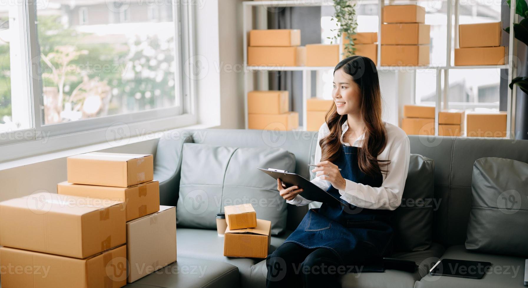 joven asiático negocio propietario mujer preparar paquete o empaquetar caja y en pie cheque en línea pedidos para entregar a cliente en tableta, ordenador portátil compras en línea concepto. foto