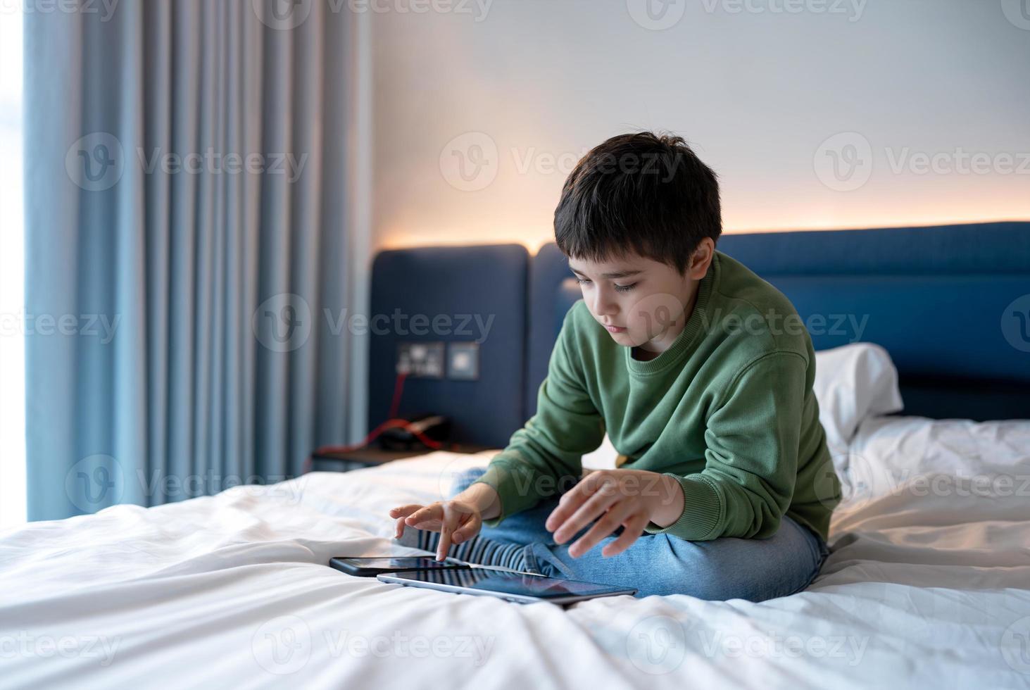 niño chico utilizando teléfono inteligente mensajes de texto a amigo, niño jugando juego en tableta mientras sentado en un cama. colegio niño utilizando móvil teléfono para deberes, niños con tecnología ,Internet, en línea educación concepto foto