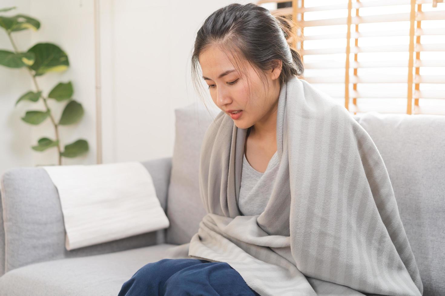 enfermo, influenza asiático joven mujer, niña dolor de cabeza tener un fiebre, gripe y cubrir frazada, sensación enfermedad sentado en sofá cama a hogar. salud cuidado persona en virus, COVID-19. foto