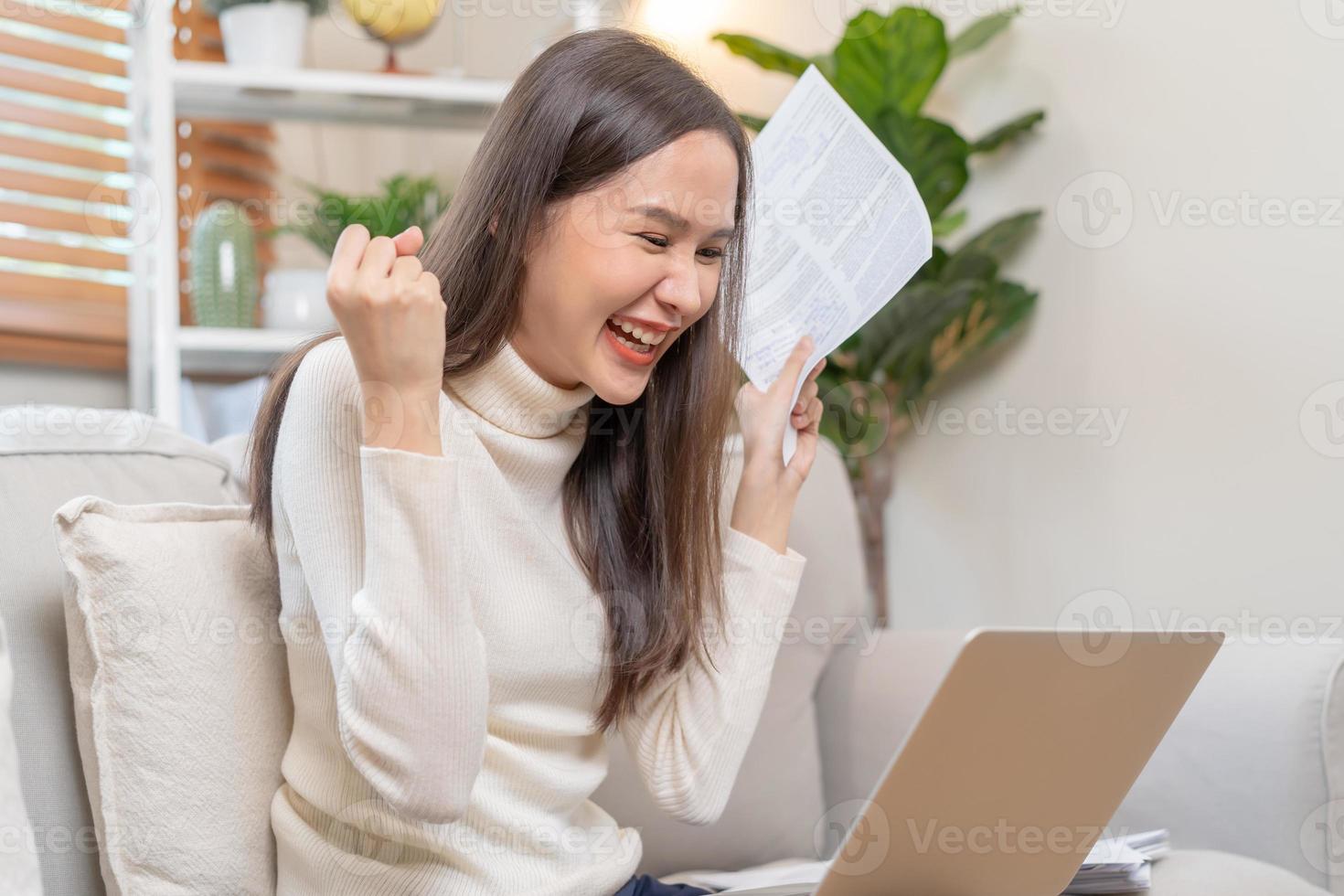 Happy excited, asian young woman holding paperwork or document getting, received job promotion, approve tax refund or loan mortgage, looking good news at laptop computer, sitting on sofa at home. photo