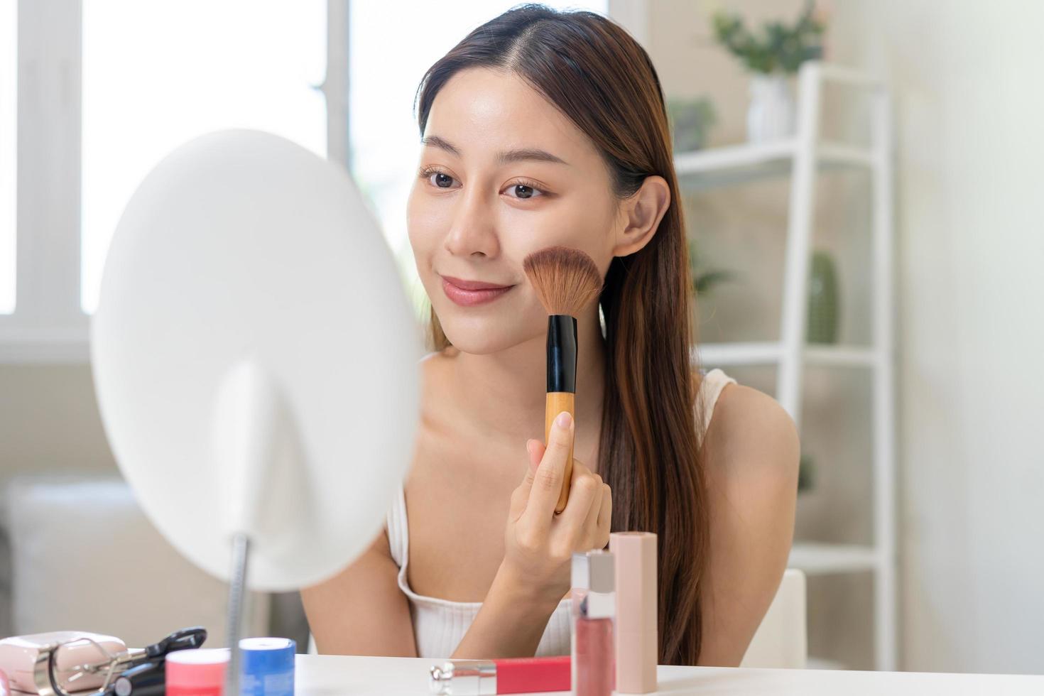 Happy beauty blogger concept, cute asian young woman, girl smile, make up face by applying brush blush powder on her cheek, looking at the mirror. People look with natural fashion style. photo