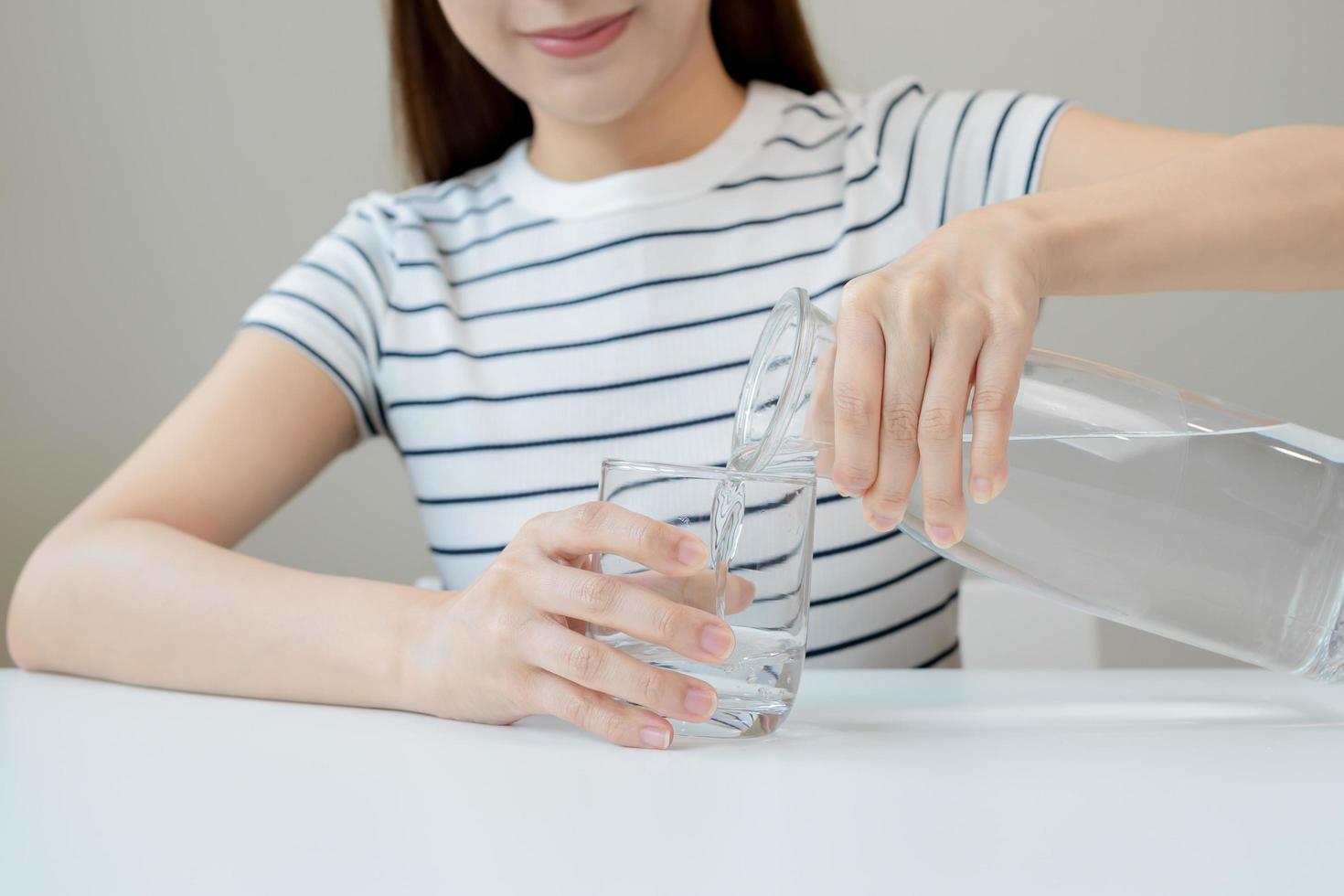 contento hermoso, sediento asiático joven mujer, niña Bebiendo, mano en tenencia, torrencial agua desde jarra dentro transparente vaso desde lanzador para hidratación de cuerpo. salud cuidado, sano estilo de vida concepto. foto