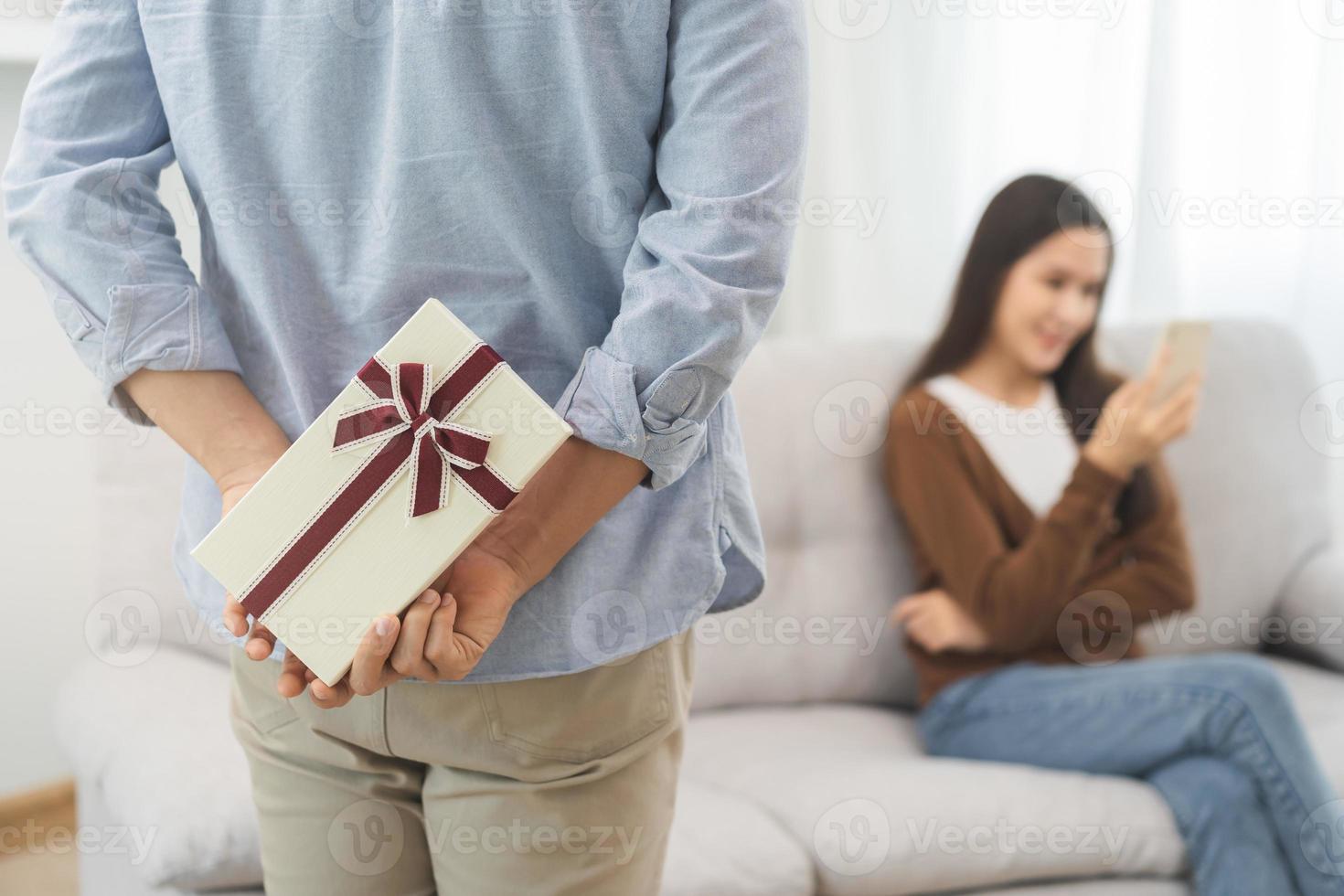 celebrando San Valentín día aniversario, relación dos asiático joven Pareja amar, novio da un regalo a Novia por esconder caja a el detrás, mujer consiguiendo presente mientras sentado en sofá a hogar. foto