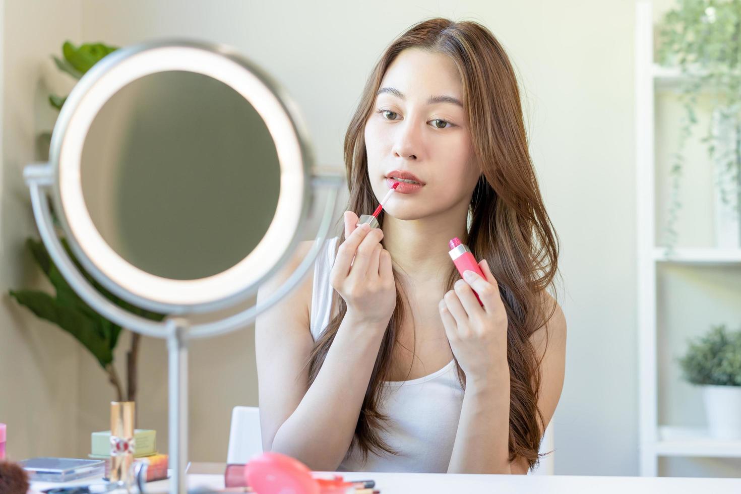 Beauty cosmetics concept, hand of asian young woman, girl make up face by applying lipstick, lips balm on her mouth, looking at the mirror at home. Female look with natural fashion style, skin care. photo