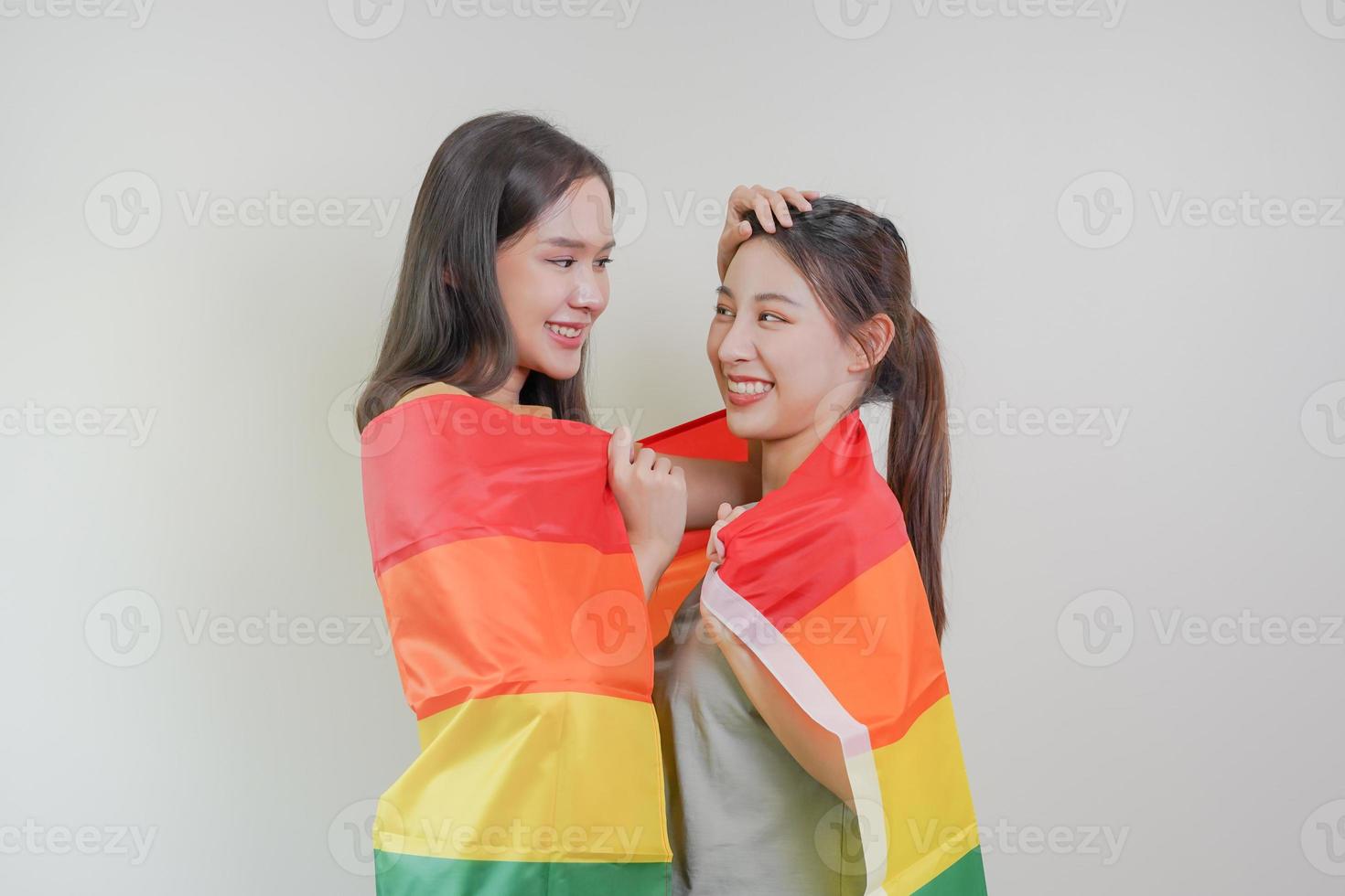 Happy lesbian, beautiful asian young two women, girl gay, couple love moment spending good time together, holding or waving lgbt rainbow, pride flag on isolated white wall background together at home. photo