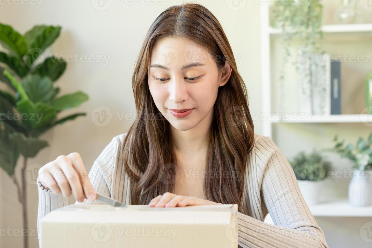 Happy asian young woman, girl customer sitting on sofa at home, hand in opening and unpacking cardboard box carton parcel after buying ordering present, shopping online, delivery service concept. photo