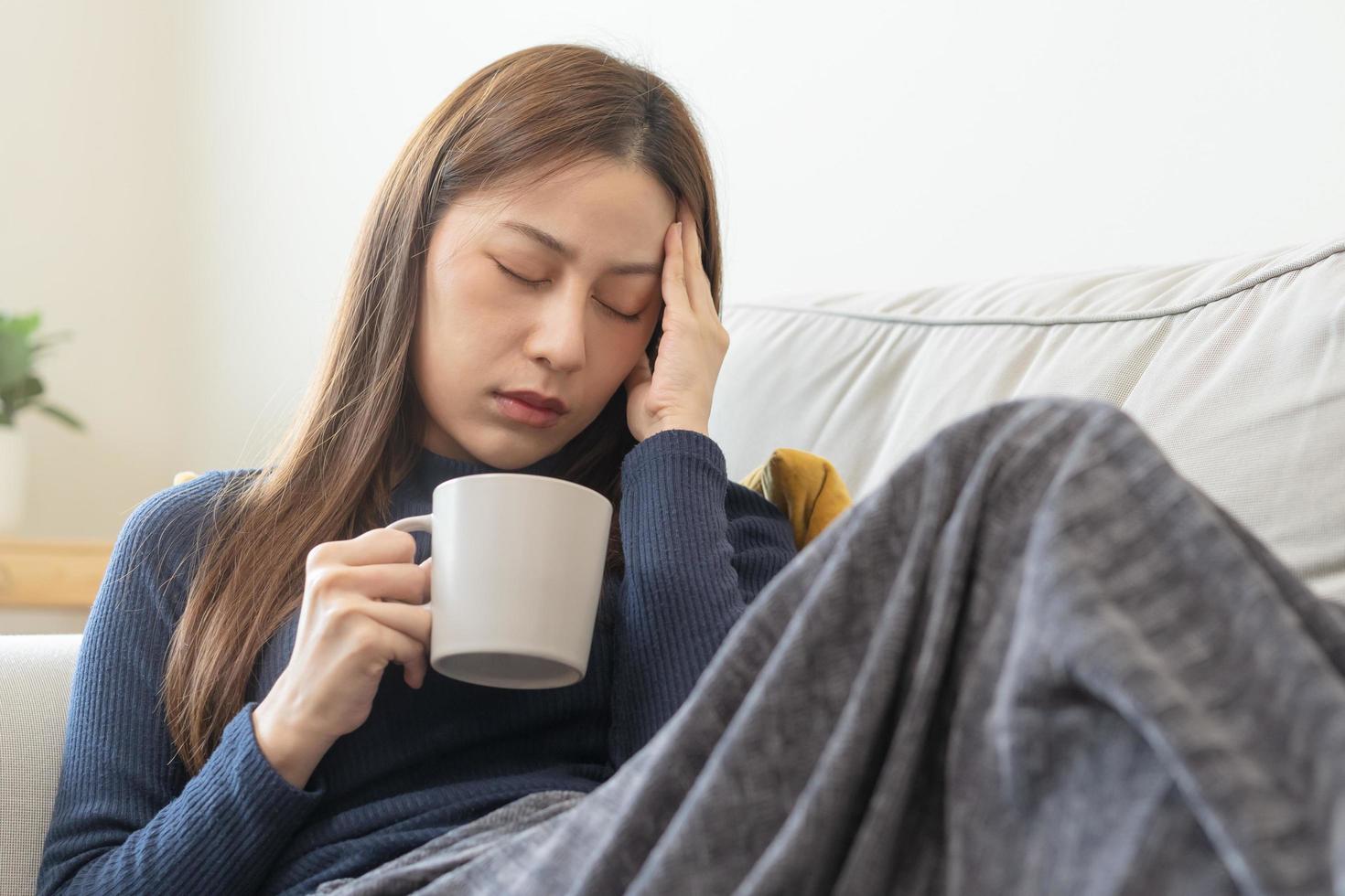 enfermo, influenza asiático joven mujer, niña dolor de cabeza tener un fiebre, gripe y cheque medida cuerpo temperatura, sensación enfermedad sentado en sofá cama a hogar. salud cuidado persona en virus, COVID-19. foto