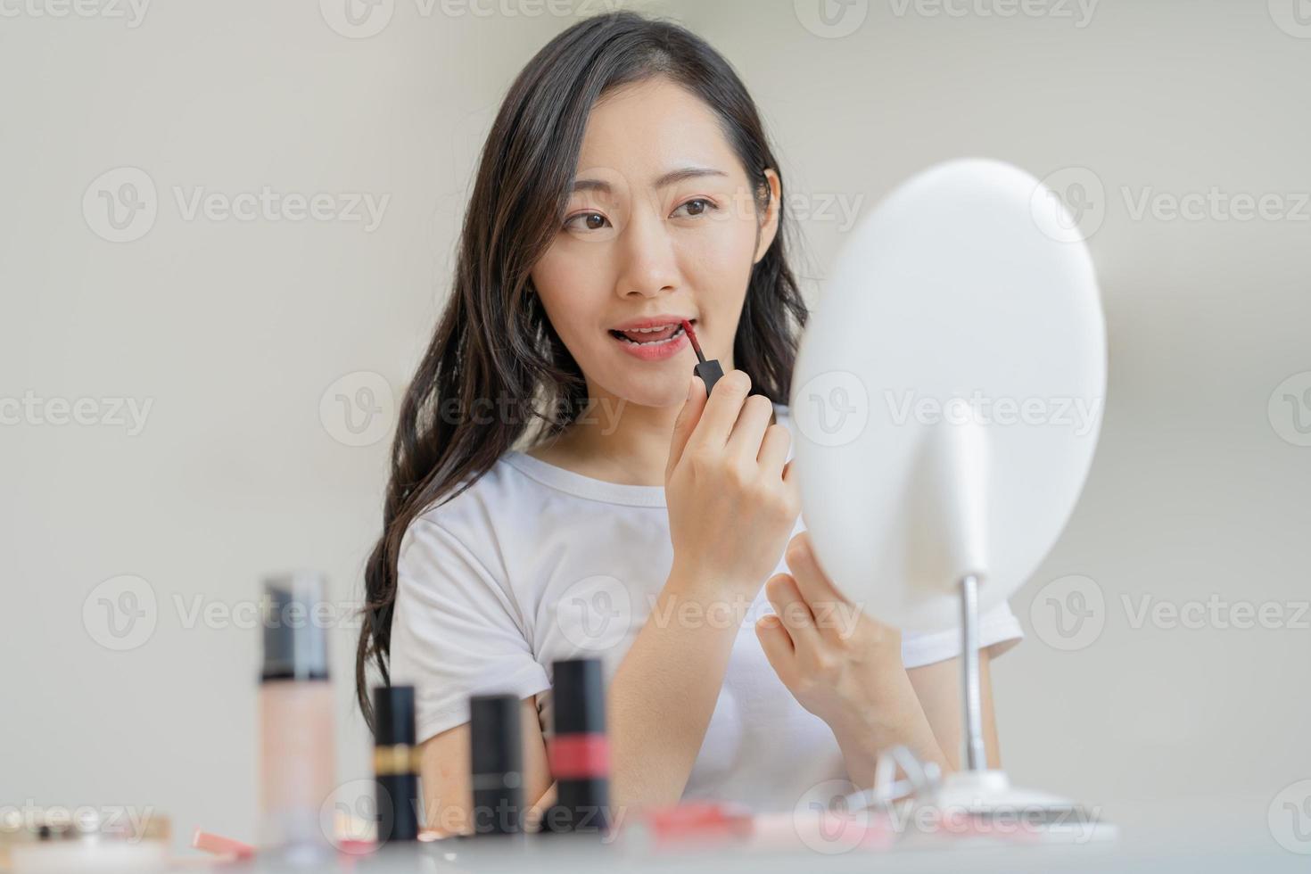 belleza productos cosméticos concepto, mano de asiático joven mujer, niña hacer arriba cara por aplicando lápiz labial, labios bálsamo en su boca, mirando a el espejo a hogar. hembra Mira con natural Moda estilo, piel cuidado. foto