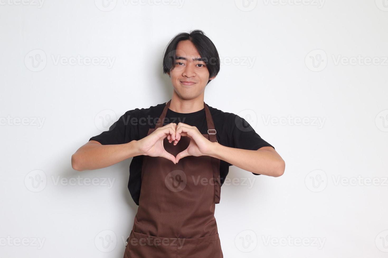 retrato de atractivo asiático barista hombre en marrón delantal habla acerca de propio sentimientos, hace corazón gesto terminado pecho, expresa simpatía y amar, sonrisas afirmativamente. aislado imagen en blanco antecedentes foto