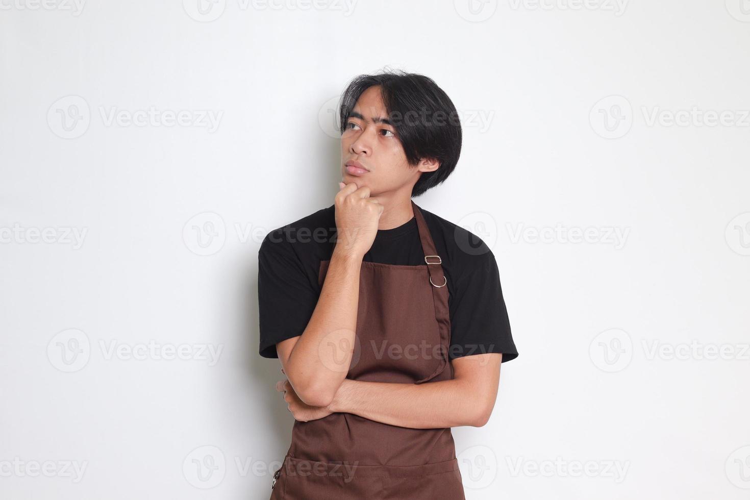 Portrait of attractive Asian barista man in brown apron thinking about question with hand on chin. Isolated image on white background photo