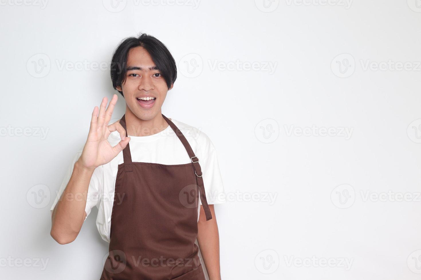 Portrait of attractive Asian barista man in brown apron showing ok hand gesture and smiling looking at camera. Advertising concept. Isolated image on white background photo
