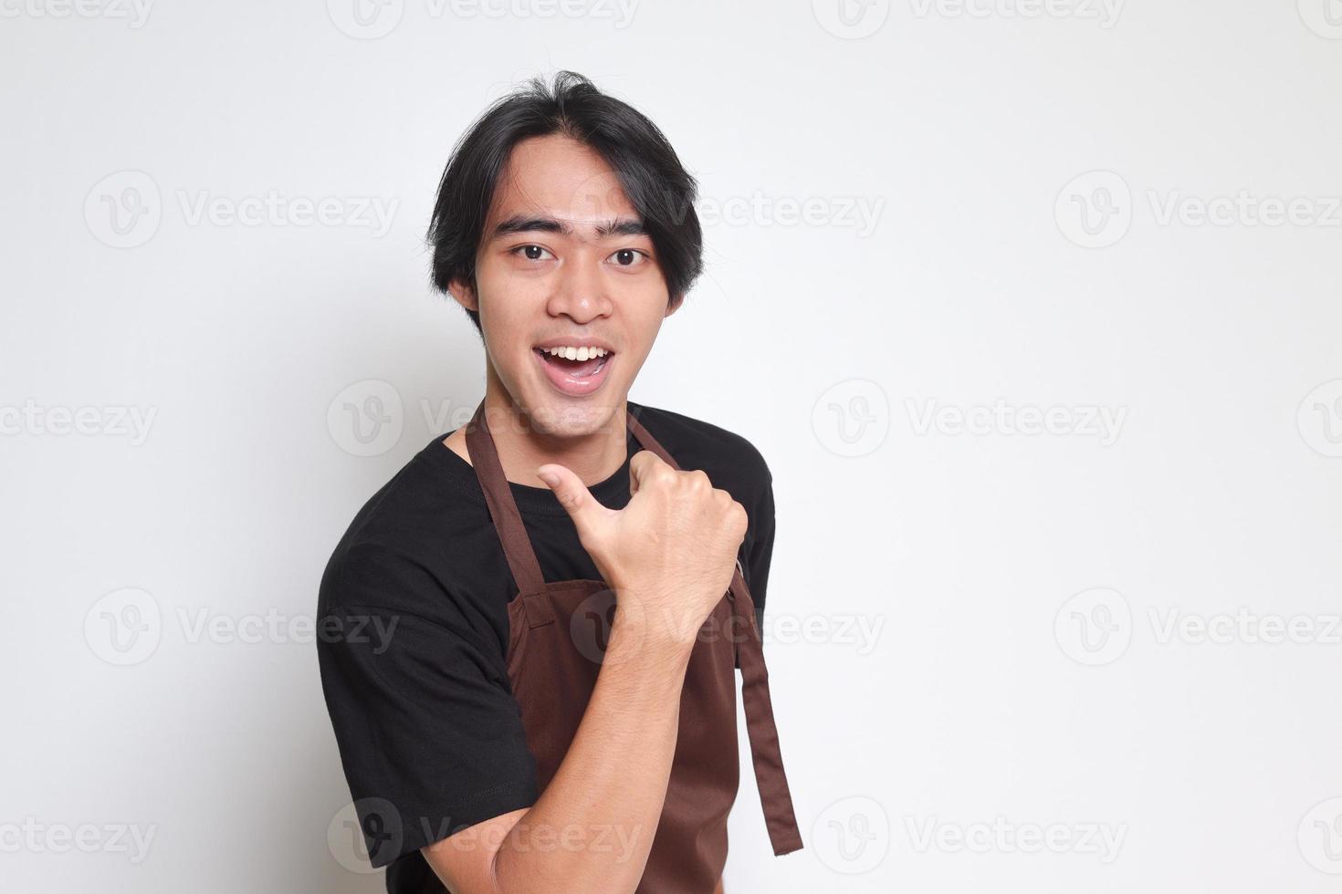 Portrait of attractive Asian barista man in brown apron with thumb pointing away on empty space. Advertising concept. Isolated image on white background photo