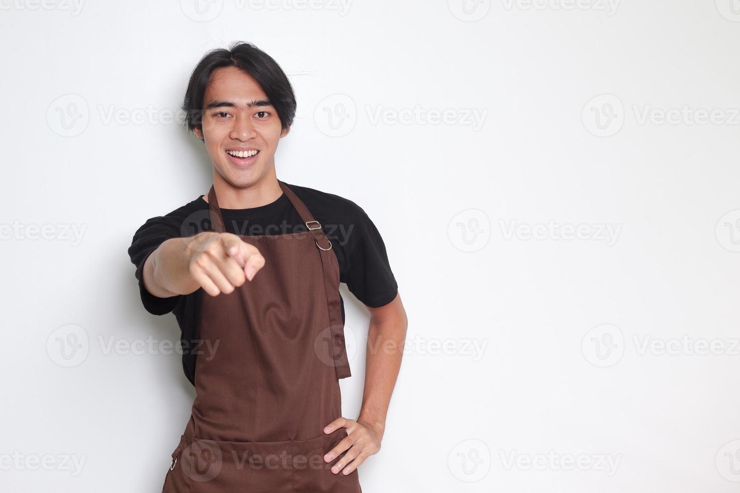 Portrait of attractive Asian barista man in brown apron pointing forward to the camera with finger. Advertising concept. Isolated image on white background photo