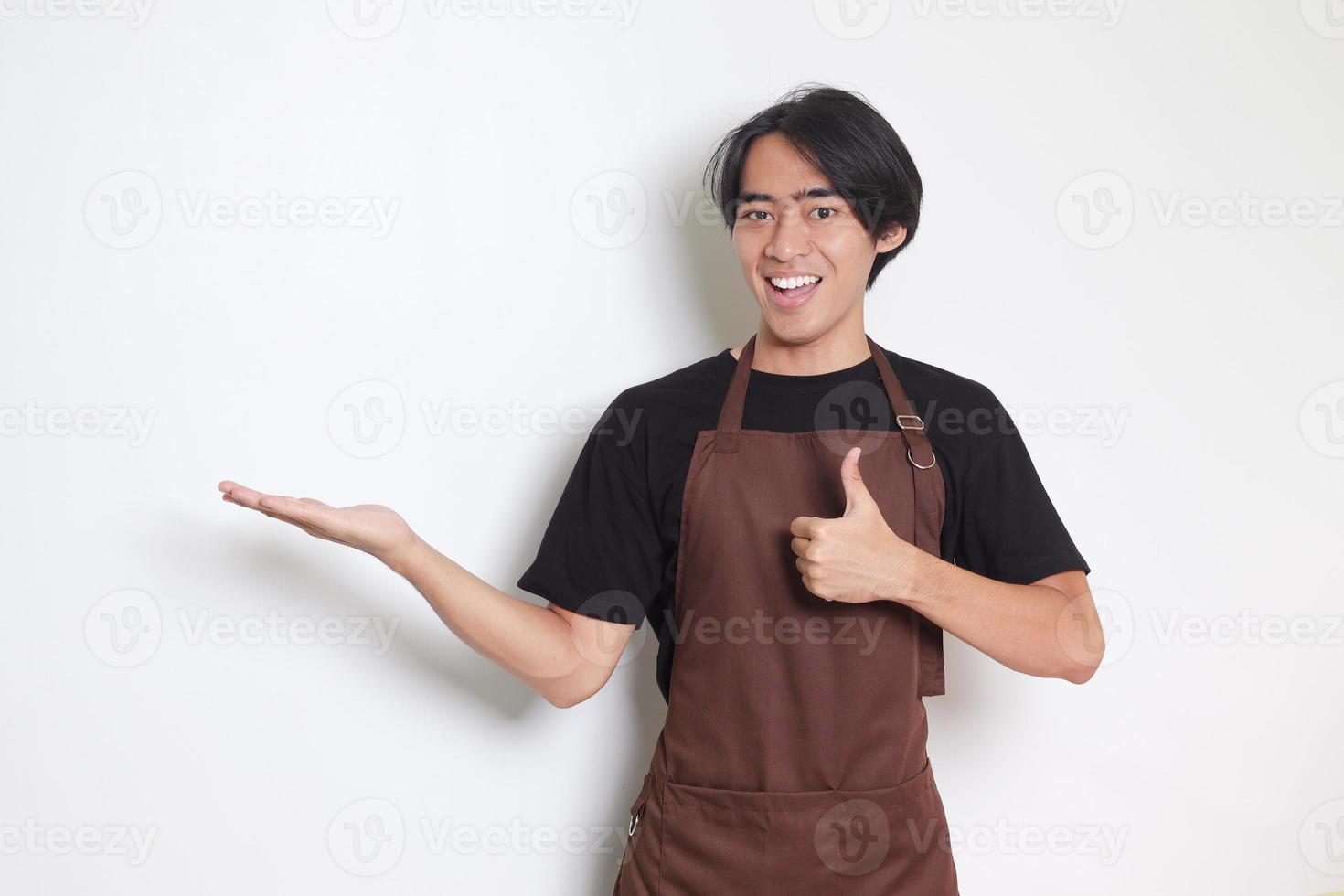 retrato de atractivo asiático barista hombre en marrón delantal demostración producto, señalando a alguna cosa con manos. publicidad concepto. aislado imagen en blanco antecedentes foto