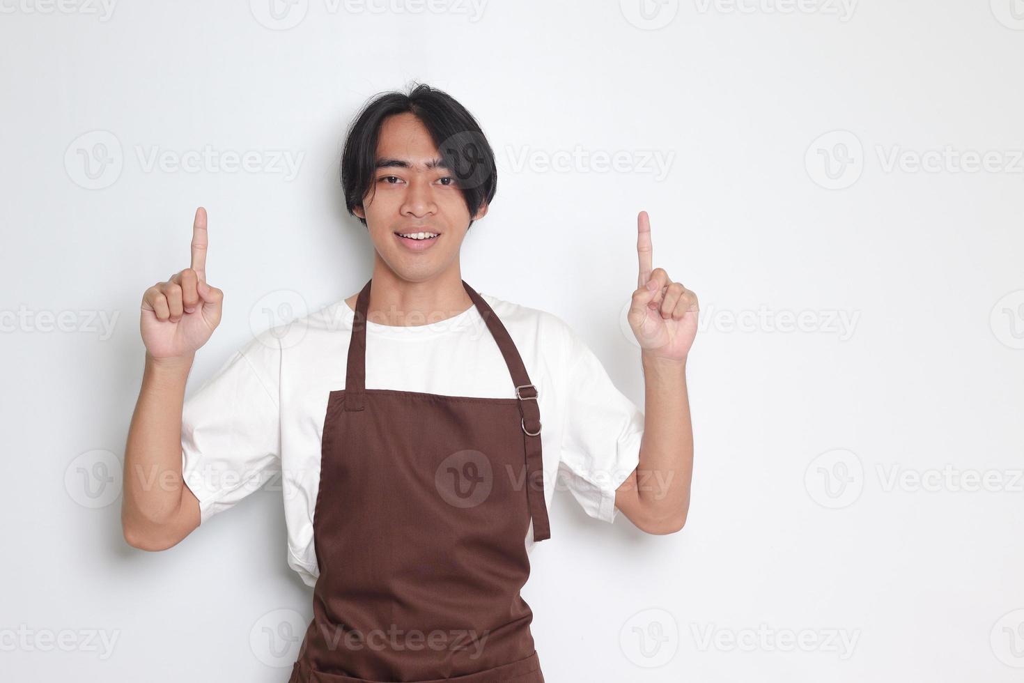 retrato de atractivo asiático barista hombre en marrón delantal demostración producto, señalando a alguna cosa con manos. publicidad concepto. aislado imagen en blanco antecedentes foto