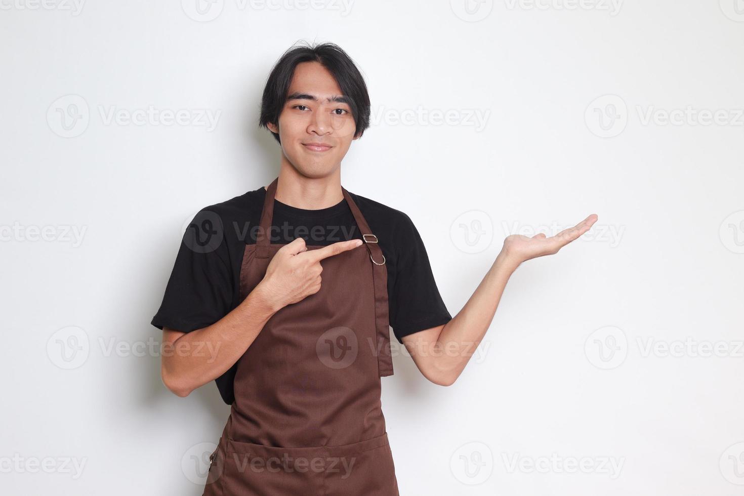 Portrait of attractive Asian barista man in brown apron showing product, pointing at something with hands. Advertising concept. Isolated image on white background photo