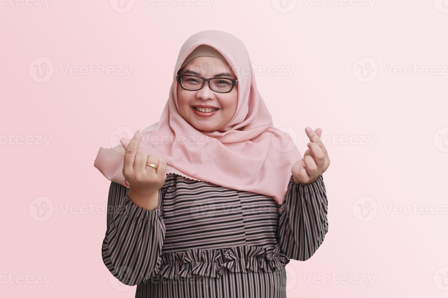 Portrait of cheerful Asian woman with hijab, showing finger sign of Korean love. Isolated image on pink background photo