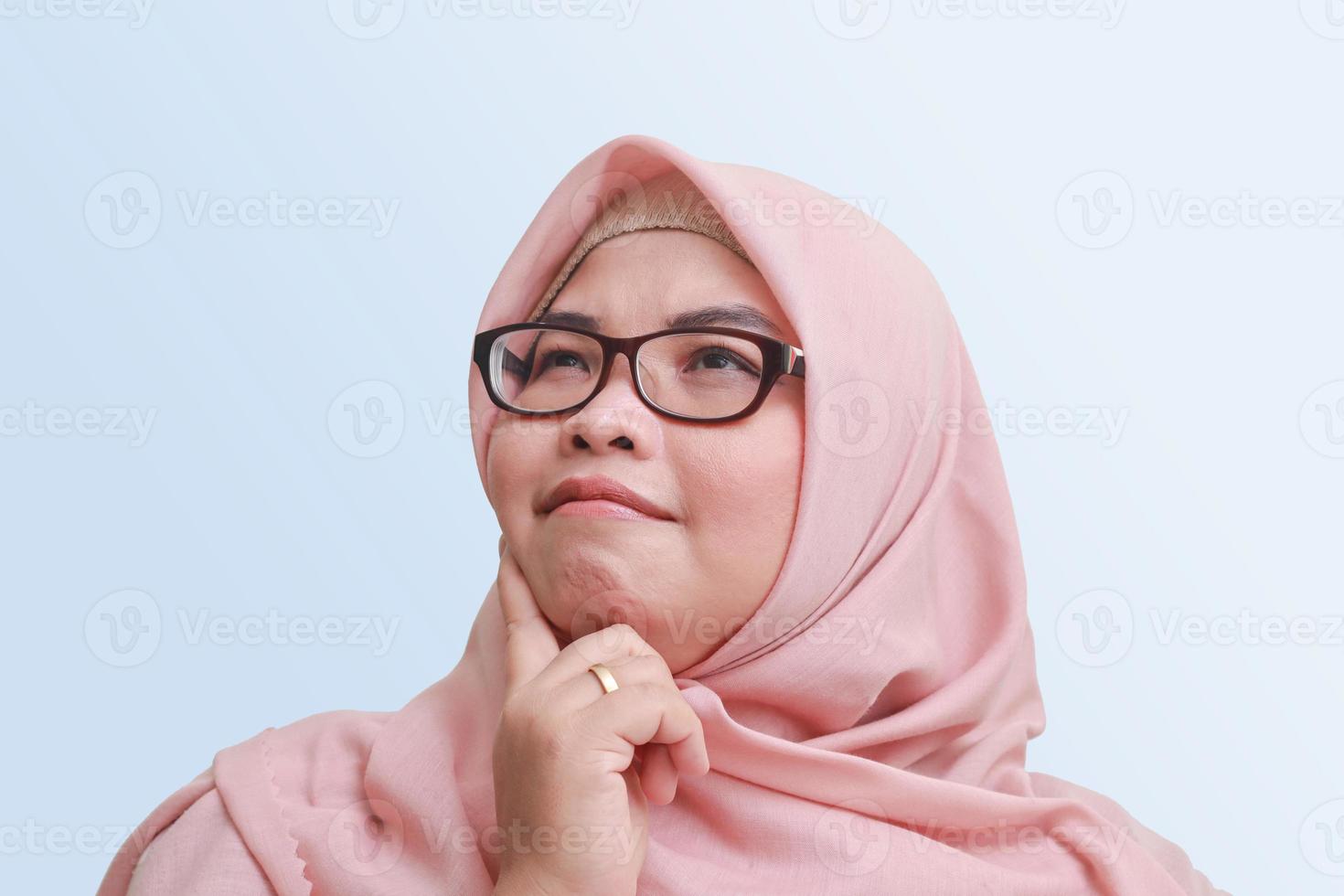 Portrait of cheerful Asian woman with hijab, standing against blue background, thinking about question with hand on chin photo