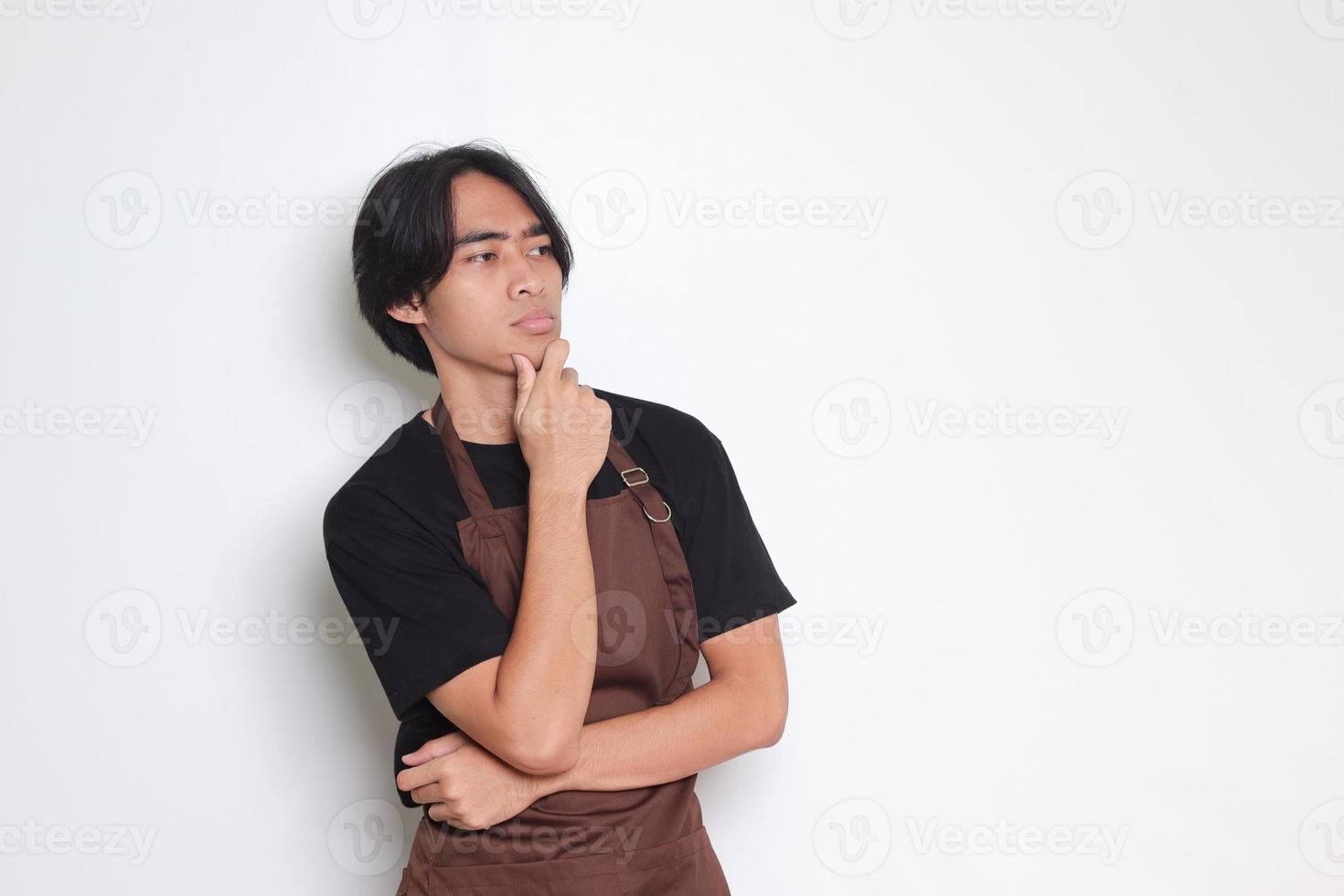 Portrait of attractive Asian barista man in brown apron thinking about question with hand on chin. Isolated image on white background photo