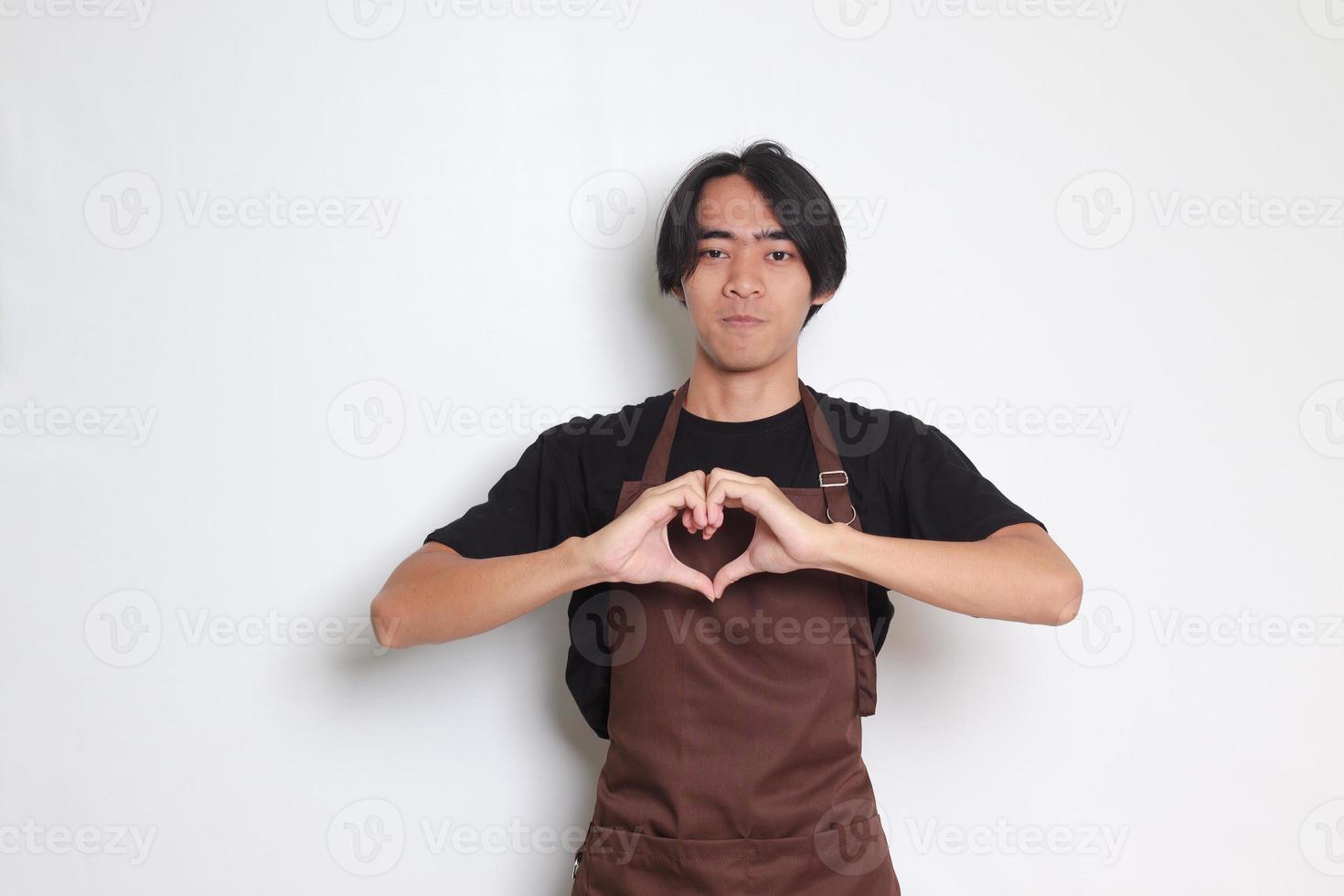 Portrait of attractive Asian barista man in brown apron speaks about own feelings, makes heart gesture over chest, expresses sympathy and love, smiles positively. Isolated image on white background photo
