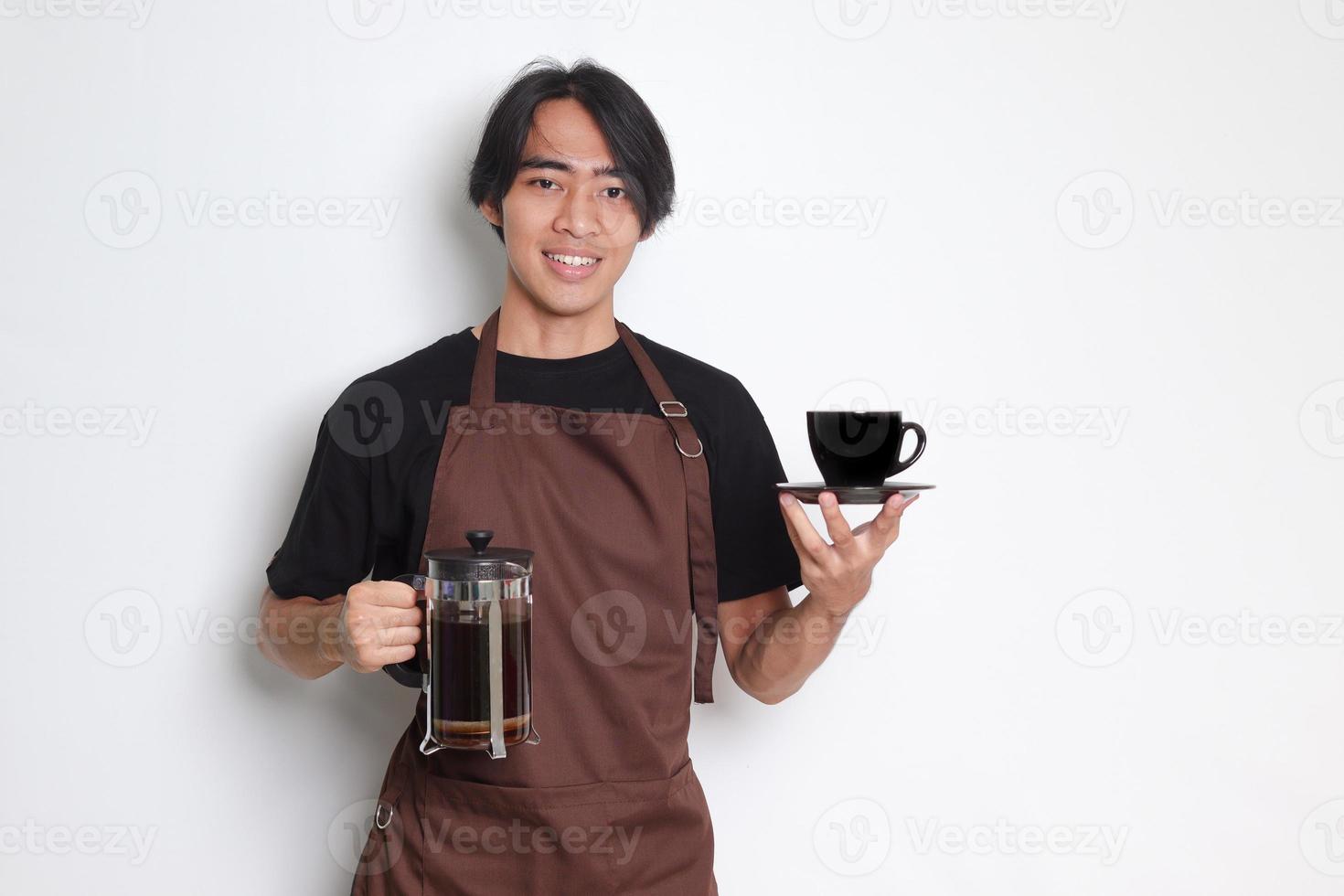 Portrait of attractive Asian barista man in brown apron showing a cup and saucer while holding french press coffee maker. Isolated image on white background photo