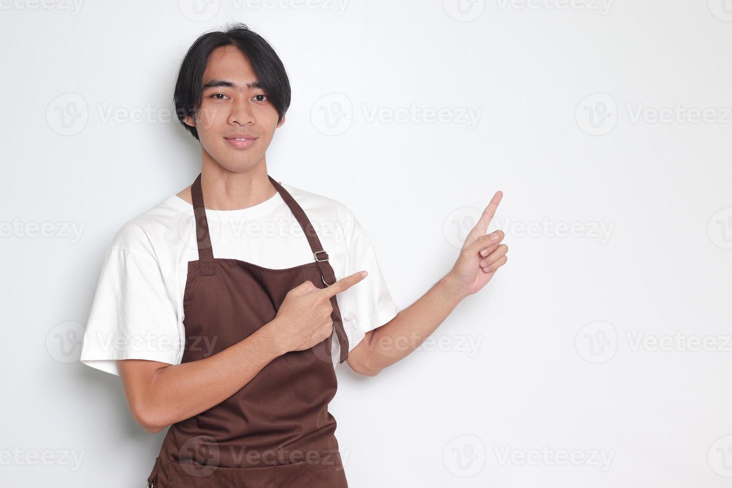 Portrait of attractive Asian barista man in brown apron showing product, pointing at something with hands. Advertising concept. Isolated image on white background photo