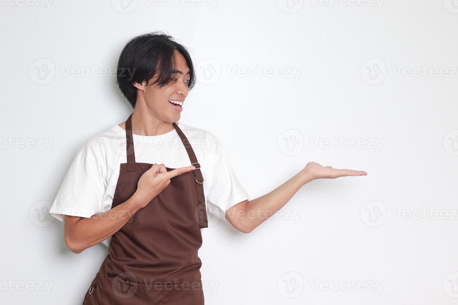 retrato de atractivo asiático barista hombre en marrón delantal demostración producto, señalando a alguna cosa con manos. publicidad concepto. aislado imagen en blanco antecedentes foto
